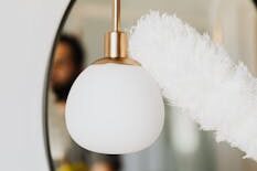 Anonymous person in casual clothes and yellow rubber glove cleaning white stylish lamp with antistatic brush dust cleaner while standing against cozy oval mirror